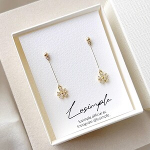 May include: A pair of gold-tone drop earrings featuring a small stud top connected by a thin chain to a flower-shaped charm set with clear stones. The earrings are displayed on a white card printed with the text Lusimple, lusimple.official.ec, Instagram @lusimple.