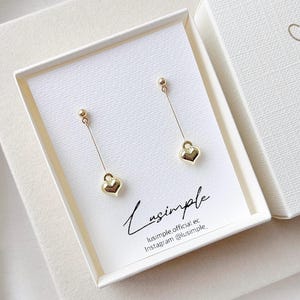 Op de afbeelding: Goudkleurige oorhangers met hartvormige bedels. De oorbellen worden gepresenteerd in een witte doos met de merknaam "Lusimple" en social media-informatie. De oorbellen liggen op een witte kaart.