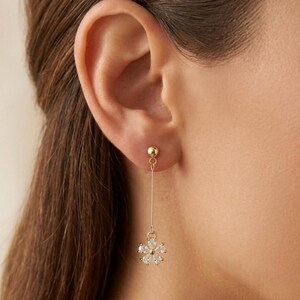 May include: A gold-tone drop earring featuring a small spherical stud at the top, a thin vertical chain, and a flower-shaped pendant at the bottom. The flower pendant is composed of five clear, pear-cut stones arranged in a circular pattern.