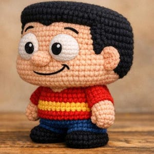 Puede incluir: Muñeco de ganchillo hecho a mano de un niño de dibujos animados con cabello negro, una cara sonriente y ojos blancos grandes. Viste una camisa roja con rayas amarillas, pantalones azules y zapatos negros. La muñeca está sobre una superficie de madera.