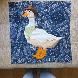 Può includere: Un blocco di tessuto quadrato per trapuntatura raffigurante un'oca composta da vari pezzi, che indossa un cappello da cowboy marrone, un fazzoletto a quadri giallo chiaro e stivali da cowboy arancioni su sfondo blu scuro con motivo paisley.