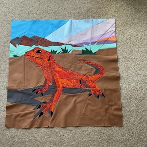 Puede incluir: Una obra de arte textil que representa un lagarto naranja brillante en un paisaje desértico. El lagarto es el punto focal, con un patchwork detallado. El fondo incluye un cielo azul, montañas y plantas del desierto.