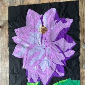 May include: A handmade quilt depicting a large, stylised purple flower on a black backdrop. The flower is rendered in various shades of purple and lavender, with a brown centre and green leaves. The quilt is square and constructed from fabric squares.