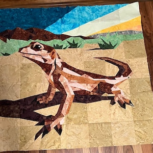 Puede incluir: Un edredón colorido con un diseño de lagarto. El lagarto es marrón y beige, con un patrón de patchwork detallado. El fondo representa un paisaje desértico con tonos azules, verdes y amarillos. El edredón es cuadrado y parece hecho a mano.