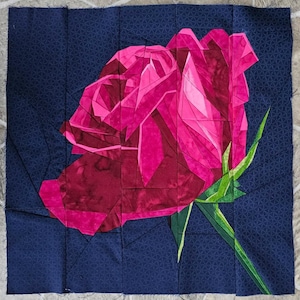 May include: A fabric art piece featuring a stylized rose in shades of pink and red against a dark blue background. The rose is constructed from geometric shapes, with green stems and leaves. The background has a subtle geometric pattern.