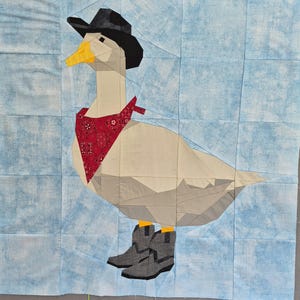 May include: A fabric quilt block featuring a pieced goose wearing a black cowboy hat, a red bandana with a white paisley pattern, and grey cowboy boots. The goose is light beige with a yellow beak, set against a light blue mottled background.