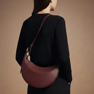May include: A burgundy crescent-shaped shoulder bag with a long strap. The bag is made of smooth leather and has a minimalist design. The person is wearing a black long-sleeve shirt and black pants.