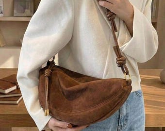 May include: A crescent-shaped brown suede shoulder bag featuring a knotted strap design with long, dangling tassel accents on the side. The bag is worn over a white blazer and light blue denim jeans, showcasing a soft, textured material.