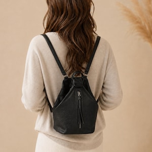 May include: A black leather backpack with a triangular silhouette, featuring a central vertical zippered pocket with a long leather pull tab, a top handle, and two adjustable shoulder straps, worn over a beige long-sleeved knit sweater.