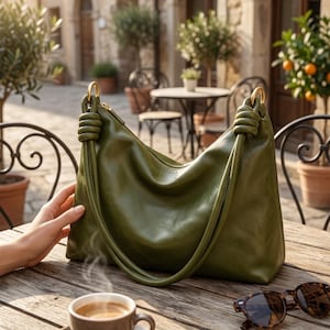 May include: An olive green leather handbag with gold-tone handles. The bag features a knotted handle and a zip closure. A cup of coffee and sunglasses sit on a wooden table. A fashionable accessory.