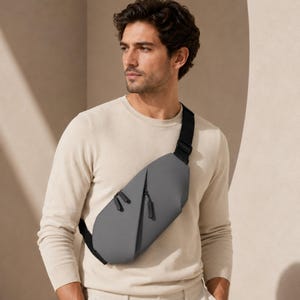 May include: A sleek, matte grey crossbody sling bag featuring a structured, minimalist design with two diagonal zippered compartments. The bag includes a wide, adjustable black strap that rests across the chest, providing a secure and functional fit.