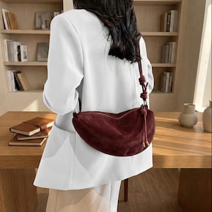 K&ouml;nnte beinhalten: Eine bordeauxfarbene Wildleder-Umh&auml;ngetasche in Halbmondform. Die Tasche hat einen geknoteten Riemen und goldfarbene Beschl&auml;ge. Die Person tr&auml;gt einen wei&szlig;en Blazer und eine beige Hose.