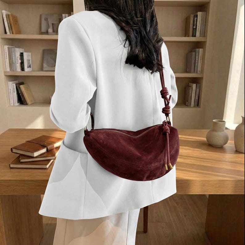 K&ouml;nnte beinhalten: Eine bordeauxfarbene Wildleder-Umh&auml;ngetasche in Halbmondform. Die Tasche hat einen geknoteten Riemen und goldfarbene Beschl&auml;ge. Die Person tr&auml;gt einen wei&szlig;en Blazer und eine beige Hose.