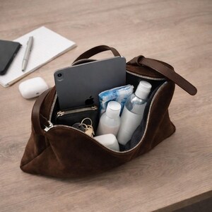 May include: A brown suede travel bag with a zipper, handle, and strap. The bag is open, revealing a tablet, small black wallet, keys, toiletries, and a small towel. A pen, notepad, phone, and wireless earbuds are on the wooden surface.