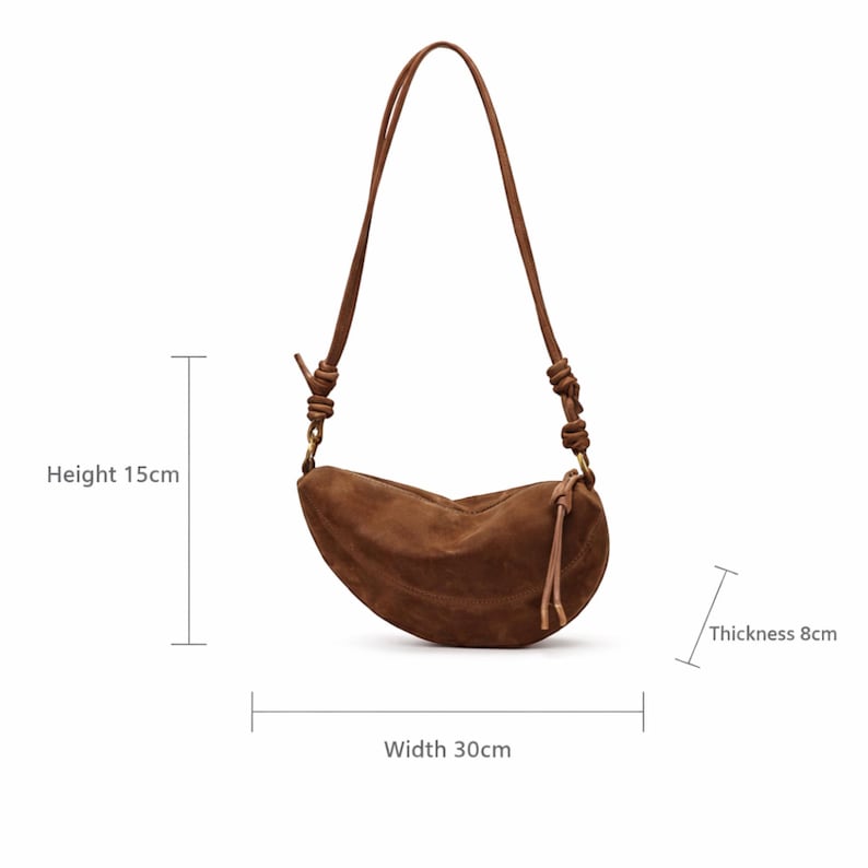 K&ouml;nnte beinhalten: Braune Wildleder-Umh&auml;ngetasche in Halbmondform. Die Tasche ist 15 cm hoch, 30 cm breit und 8 cm dick. Sie hat einen langen, verstellbaren Riemen und eine Quaste. Minimalistisches Design.