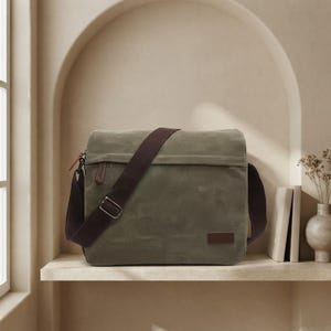 Peut inclure: Un sac messager rectangulaire en toile vert olive doté d'une bandoulière réglable marron foncé, d'un rabat supérieur, d'une petite tirette de fermeture éclair rouge sur le côté et d'un écusson rectangulaire marron foncé en bas à droite.