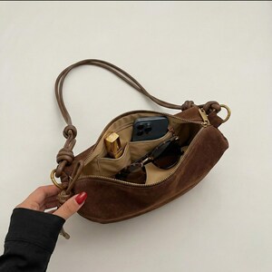 K&ouml;nnte beinhalten: Eine braune Wildleder-Umh&auml;ngetasche mit Rei&szlig;verschluss, die ein beiges Innenfutter freilegt. Im Inneren befinden sich ein Smartphone, eine Sonnenbrille und ein Lippenstift. Die Tasche hat einen langen Riemen und goldfarbene Beschl&auml;ge.
