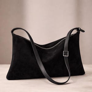May include: A black suede shoulder bag with a long, adjustable strap and silver-tone zipper. The bag has a rectangular shape with a curved top and a buckle detail on the strap. The bag is made of a soft, matte material.