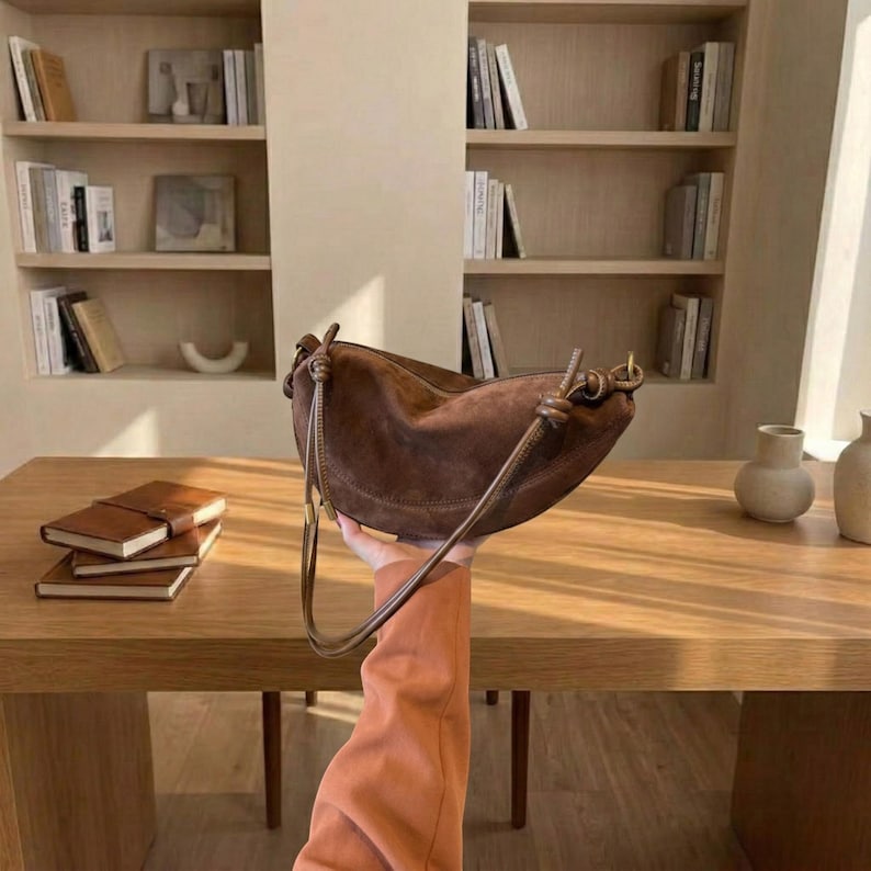 K&ouml;nnte beinhalten: Eine braune Wildleder-Umh&auml;ngetasche mit geschwungener Form und langem Riemen. Die Tasche wird vor einem Holztisch hochgehalten. Die Tasche hat ein geknotetes Detail am Riemen.