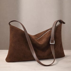 May include: A brown suede shoulder bag with a long, adjustable strap and a silver zipper. The bag has a rectangular shape with a curved top and a buckle closure on the strap. The bag is made of a soft, textured material.