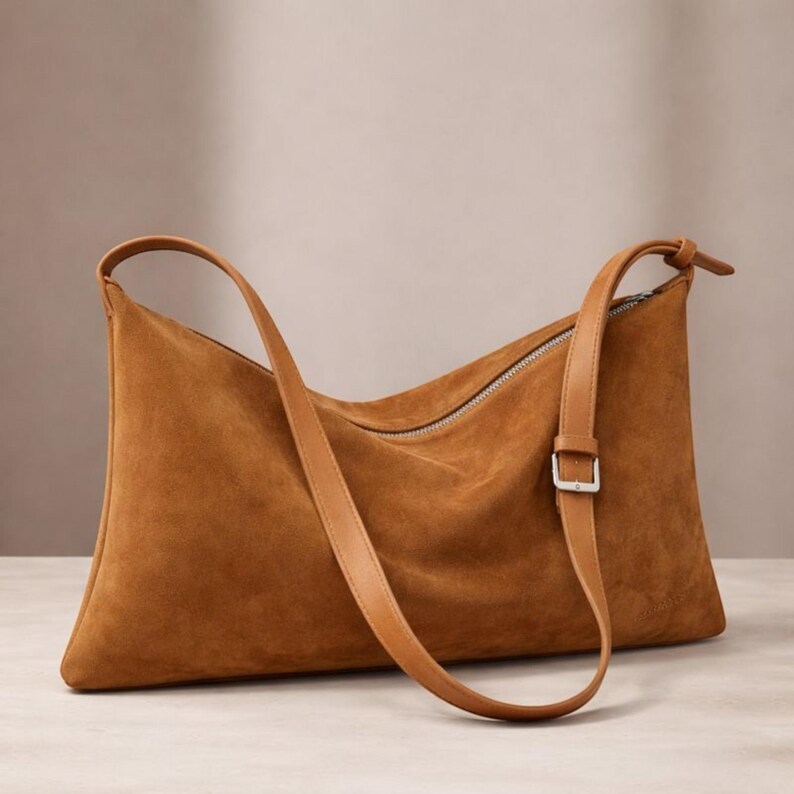 May include: A brown suede shoulder bag with a long, adjustable strap and a silver zipper closure. The bag has a rectangular shape with rounded corners and a smooth leather strap. The bag is approximately 15 inches wide and 10 inches tall.