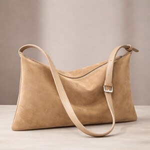 May include: A tan suede shoulder bag with a long, adjustable strap. The bag has a rectangular shape with a zipper closure and a light-colored strap. The bag is made of a soft, textured material.