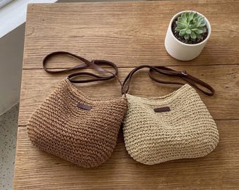May include: Two woven shoulder bags in different shades of brown and beige sit on a wooden surface. Each bag has a brown leather strap and a small, rectangular leather patch. A small potted succulent is in the background.