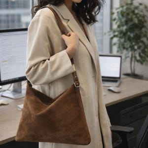 May include: A brown suede shoulder bag with a long strap. The bag has a triangular shape and is being carried over the shoulder. The strap is a darker brown and has a silver buckle. The person is wearing a beige blazer.