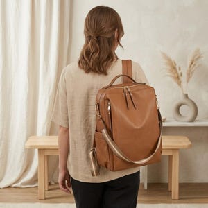 以下が含まれることがあります： ゴールドカラーのジッパーと金具が付いた、茶色の革製バックパック。バッグにはトップハンドルと調節可能なショルダーストラップがあり、そのうちの1つはストライプです。バックパックは人が着用しています。