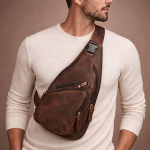 Puede incluir: Un bolso bandolera de cuero marrón con múltiples compartimentos con cremallera y un cierre de hebilla negro. El bolso se lleva cruzado sobre el pecho y el hombro. El cuero tiene un aspecto envejecido.