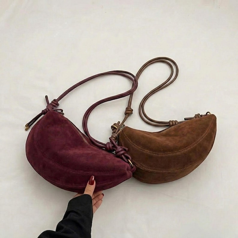 K&ouml;nnte beinhalten: Zwei halbmondf&ouml;rmige Umh&auml;ngetaschen aus Wildleder. Eine ist tief burgunderfarben, die andere warmbraun. Beide Taschen haben lange, d&uuml;nne Riemen und ein geknotetes Detail, wo der Riemen auf die Tasche trifft. Die Taschen sind vor wei&szlig;em Hintergrund.