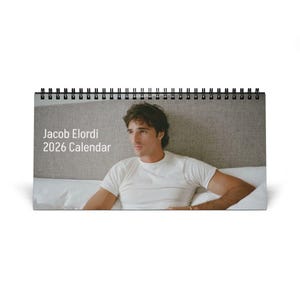 May include: A desk calendar featuring a photograph of Jacob Elordi. The calendar is spiral-bound at the top and displays the text "Jacob Elordi 2026 CALENDAR" in white. The image shows Elordi wearing a white t-shirt.