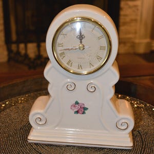 May include: A cream-colored ceramic mantel clock with a gold-rimmed circular face featuring black Roman numerals and ornate hands. The base has scroll-like accents and a small floral decal of pink roses. The word QUARTZ is printed on the dial.