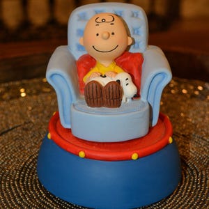 May include: A figurine of a bald character with a yellow shirt sitting in a light blue tufted armchair, holding a small white and black dog. The chair rests on a circular base featuring a red top rim with yellow dots and a solid blue side.