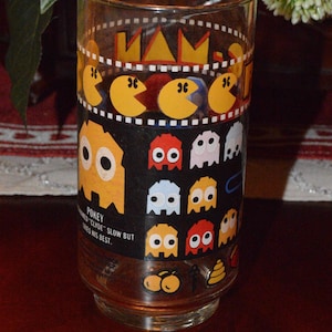 May include: A clear glass featuring a Pac-Man design. The glass has a black band with Pac-Man characters and ghosts. The text "POKEY" is visible. The glass is cylindrical and sits on a dark surface.
