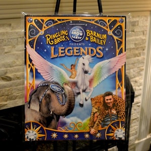 Könnte beinhalten: Ein gerahmtes Poster für die Show "Legends" von Ringling Bros. und Barnum & Bailey. Das Poster zeigt einen blauen Hintergrund mit einem weißen Pegasus, einem Elefanten und einem Mann mit einem Leoparden. Der Text enthält "The Greatest Show on Earth".
