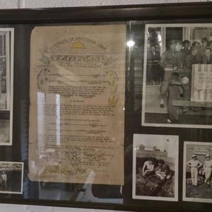 May include: A framed collection of vintage black and white photographs and documents. The display includes images of people, vehicles, and a large document with ornate borders. The photographs depict scenes from what appears to be a historical event or era.