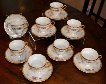 May include: A collection of vintage teacups and saucers. The cups and saucers are white with gold trim and feature a floral design with pink and purple roses. The set is arranged on a dark wooden surface.