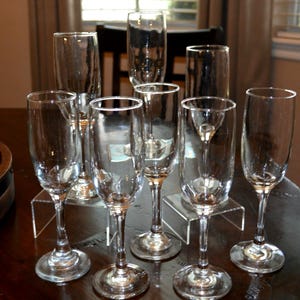 May include: A collection of clear champagne flutes and glasses arranged on a dark wooden table. The glasses vary in shape and size, with long stems and rounded bases. The background includes a window and a dark chair.