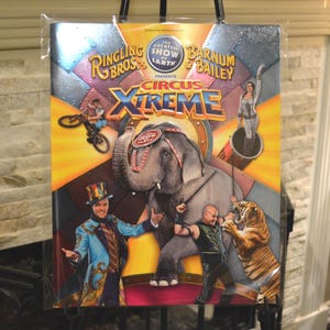 May include: A colorful poster for Ringling Bros. and Barnum & Bailey Circus Xtreme. The poster features an elephant, a tiger, performers, and the show's logo. The background has a metallic, geometric design.