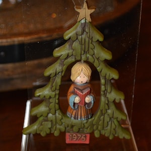 May include: A vintage Christmas ornament featuring a carved wooden figure of a child holding a book, framed by a green Christmas tree. The ornament has a gold star at the top and the year "1976" is displayed at the bottom.