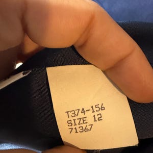 May include: A close-up of a clothing label with the text "T374-156 SIZE 12 71367". The label is cream-coloured and attached to a dark blue fabric. The background is a lighter shade of blue.