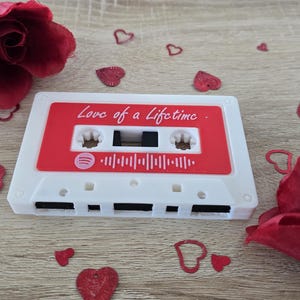 May include: A white cassette tape with a red label that reads "Love of a Lifetime" and a Spotify logo. The tape is surrounded by red rose petals and heart-shaped confetti on a wooden surface.