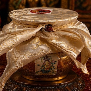 May include: A golden, ornate vessel with a decorative lid, draped with a flowing, patterned fabric. The fabric is a creamy gold color with intricate gold detailing. The vessel sits on a detailed, dark-toned base, suggesting a ceremonial or religious item.