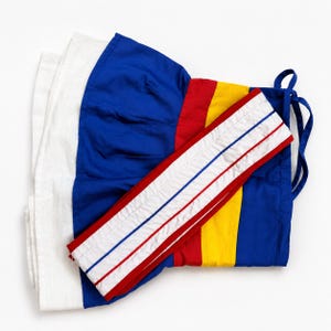 May include: A wrap-around skirt featuring vertical color-blocked panels in royal blue, bright yellow, and red, with a white textured side panel. The waistband is white with two thin horizontal red and blue stripes and includes blue fabric ties.