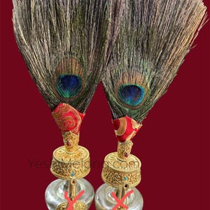 May include: Two decorative items with peacock feathers, red fabric, and gold-coloured bases. The feathers show blue, green, and brown colours. The bases have a clear glass bulb shape. The background is a deep red.