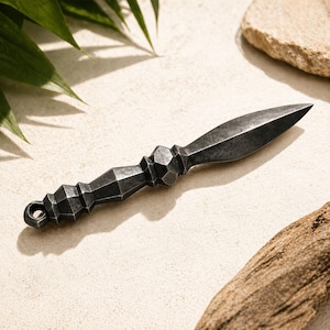 May include: A dark gray, hand-forged metal dagger with a geometric handle and a pointed blade. The dagger has a small loop at the end of the handle. The background is a neutral color with natural elements.