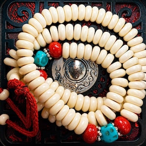 May include: Cream-colored prayer beads with red and turquoise accents. The beads are arranged in multiple strands, with a red tassel at the end. A silver-colored metal disc is in the center.