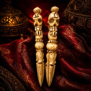 May include: Two off-white, carved bone-like hair sticks featuring a detailed skull at the top of each handle. The shafts are cylindrical with decorative bands and taper to a pointed end. The surface has a weathered, aged texture and appearance.