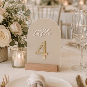 May include: An arched beige acrylic table number sign featuring the white script text "table" and a large gold reflective number "4" mounted on a rectangular wooden base. The sign is positioned upright on a table setting.
